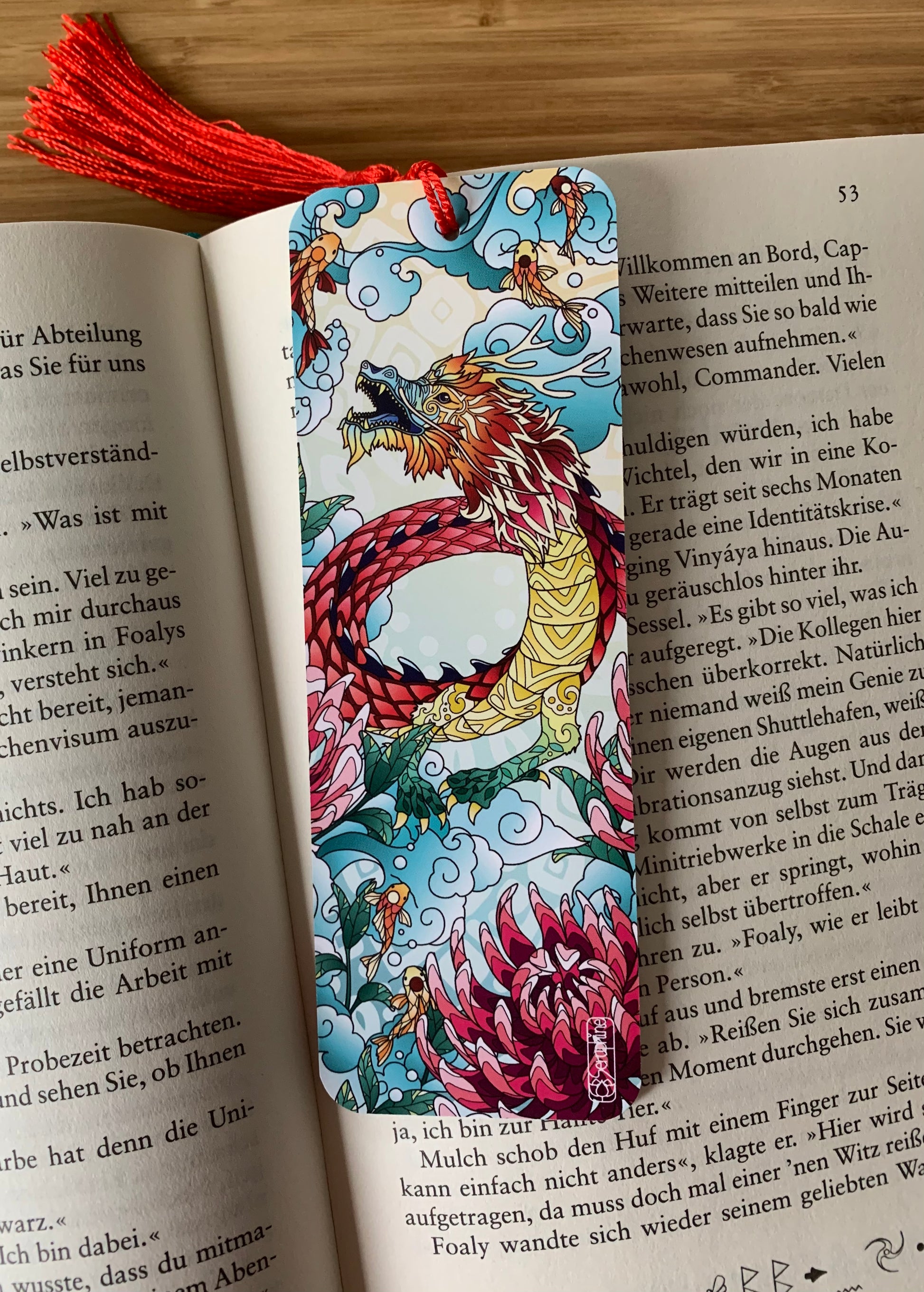 Handgemachtes Lesezeichen mit Drachenmotiv und roter Quaste in einem Buch.